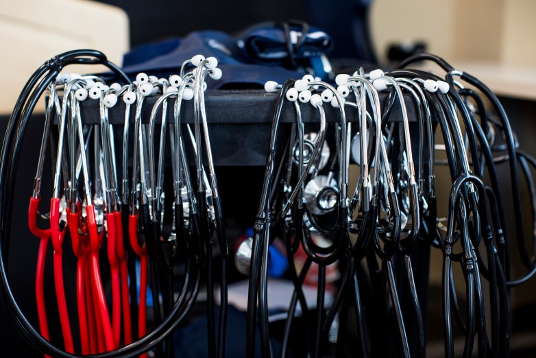 Rows of stethoscopes with black and red tubing hang from a cart beside folded blood pressure cuffs.