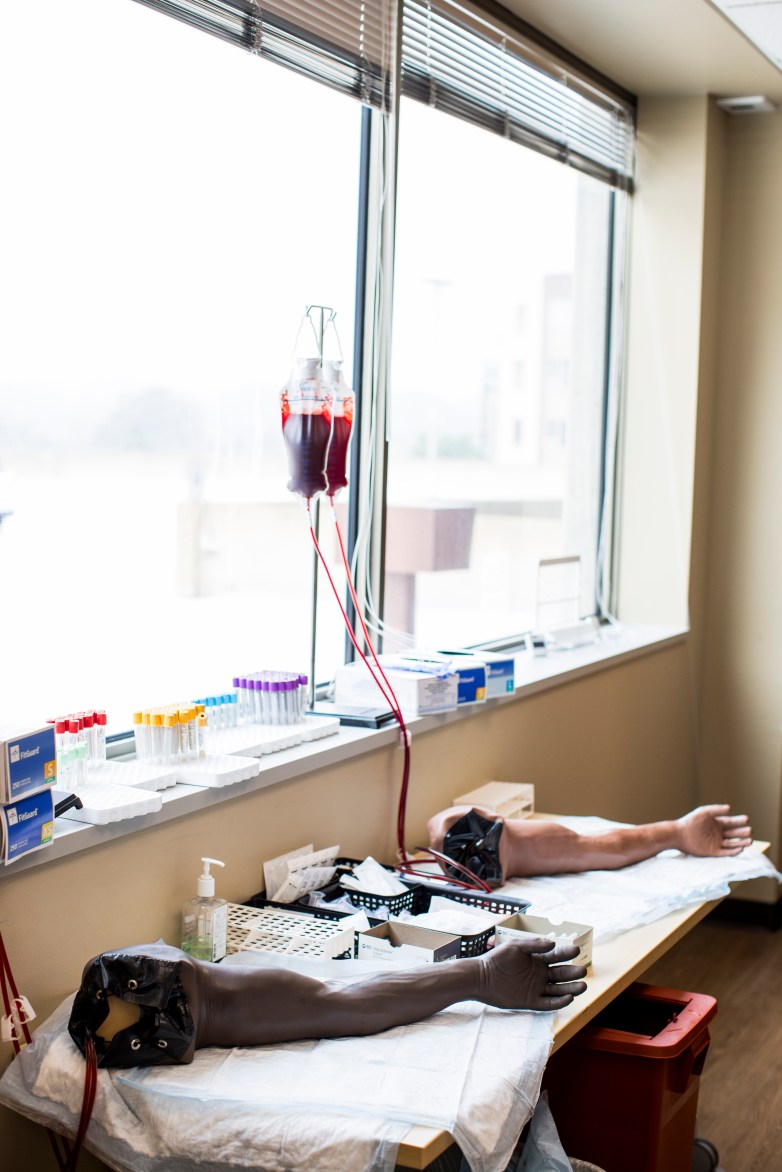 Two mannequin arms lie on tables beneath hanging blood collection bags. Blood collection tubes and medical supplies are arranged along the windowsill.