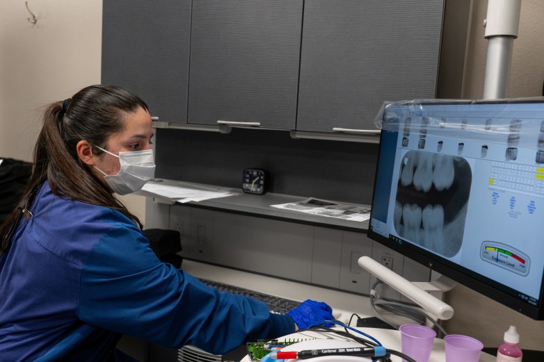 A person wearing a white face mask and blue gloves looks at a computer monitor displaying dental X-rays while seated at a workstation.