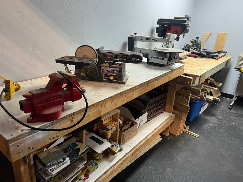A woodworking bench holds a vise, belt sander, scroll saw, drill press and other shop tools. Shelves beneath the bench store wood pieces and supplies.
