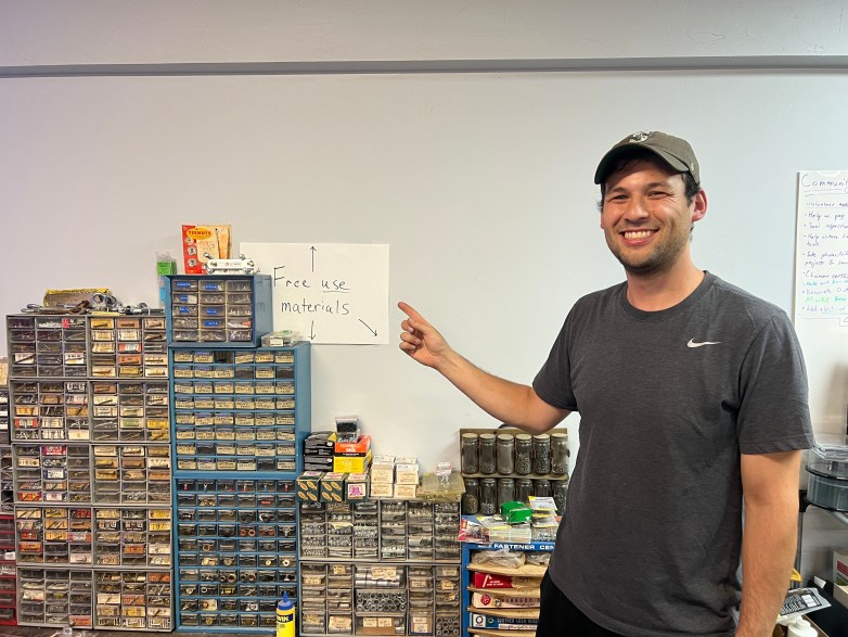 A person points to a handwritten sign reading "Free use materials" above drawers and containers filled with hardware and fasteners.