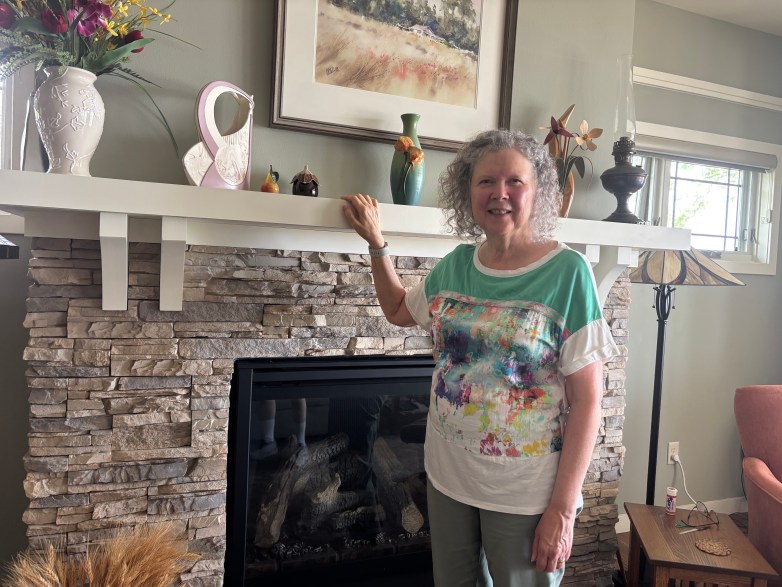 A person stands beside a stone fireplace with one hand resting on the mantel, surrounded by vases, framed artwork and decorative objects.