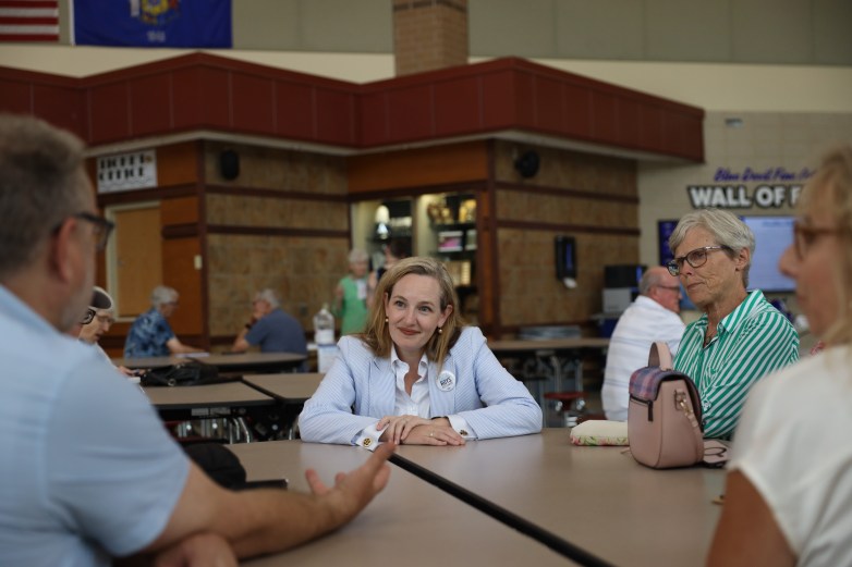 People sit around a table in a cafeteria setting. A person wearing a button reading "ROYS" sits among the group while others are seated in the background.