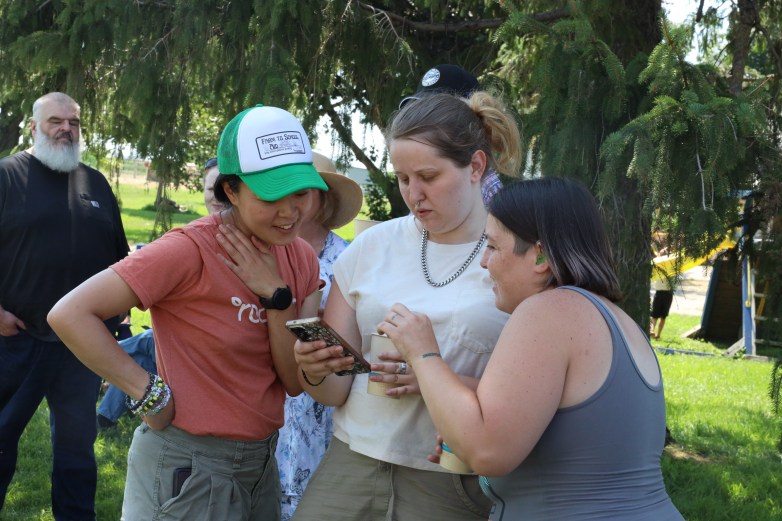 Three people stand together outdoors looking at a smartphone while others gather under trees in the background.