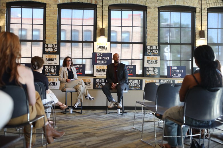 People sit facing two people indoors. Campaign signs reading "MANDELA BARNES FOR WISCONSIN," "END CITIZENS UNITED" and "CORPORATIONS ARE NOT PEOPLE" are displayed behind them next to windows.