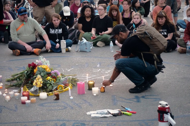 A person kneels to light a candle beside flowers, candles and yellow caution tape on pavement while people sit in a circle around the memorial.