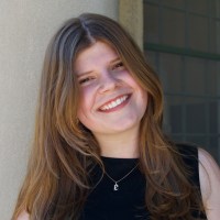 A person with long brown hair and a nose ring wears a black top and a necklace with an "E" pendant.