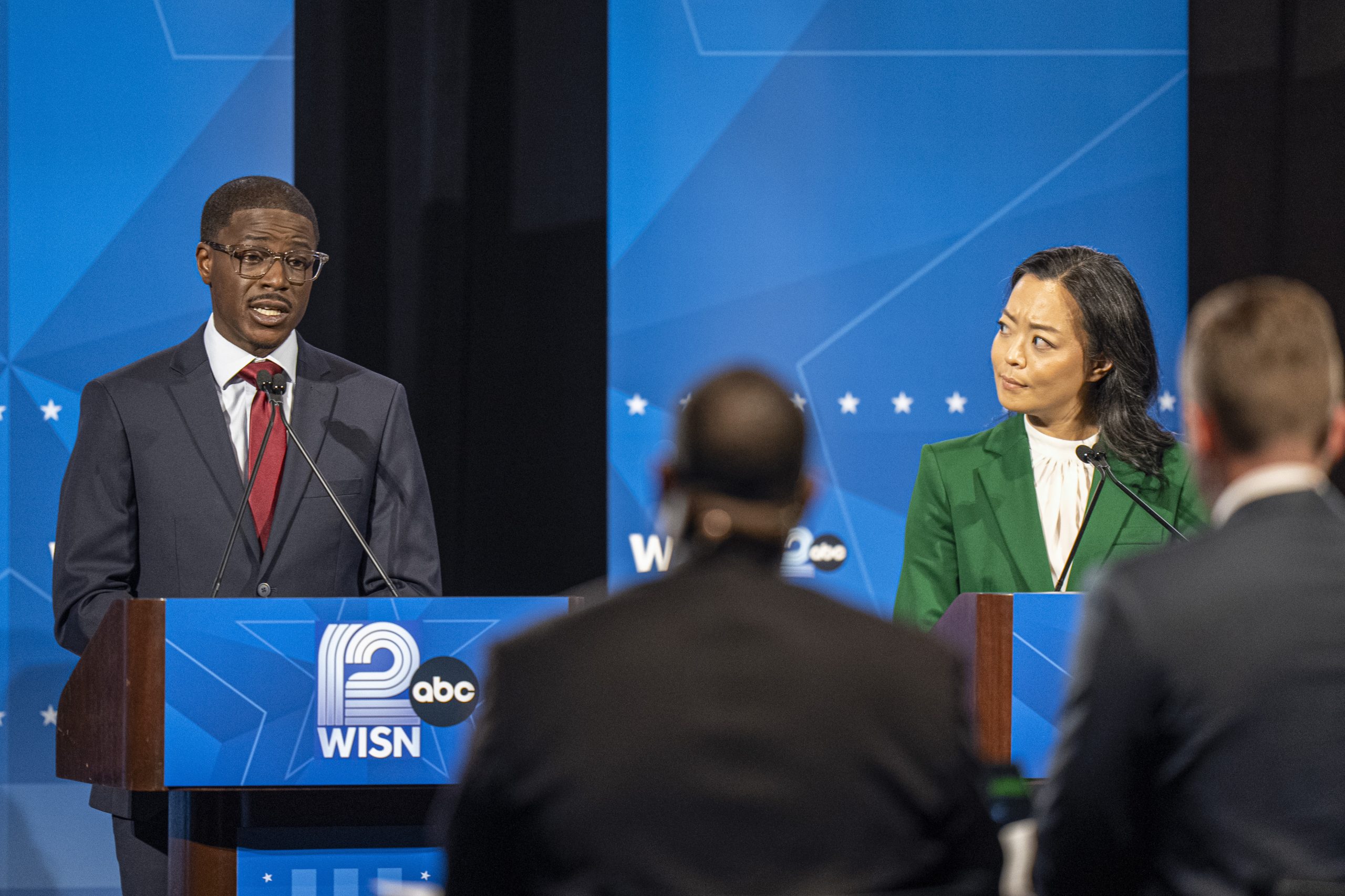 Photo: Wisconsinwatch A person in a dark suit speaks at a "WISN 12" podium while another person in a green jacket looks on, with two people seen