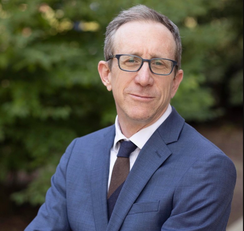 A person wearing glasses, a blue suit and a dark tie faces the camera, with green trees blurred in the background.