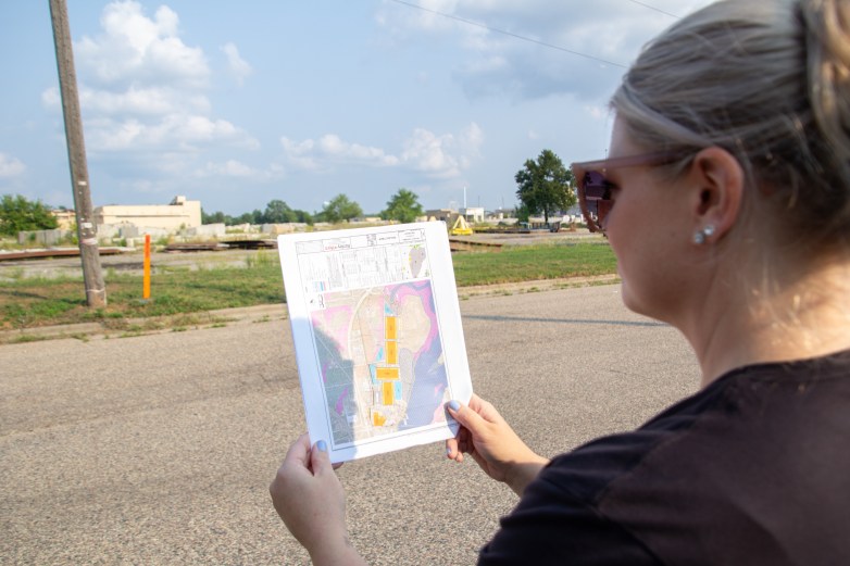 A person wearing sunglasses holds a printed map while standing across a road from an industrial area.