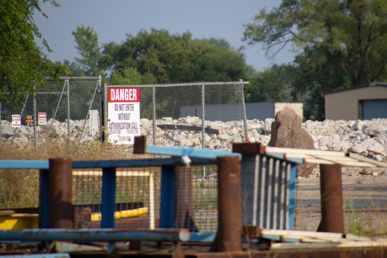 A chain-link fence is next to piles of rocks and industrial materials. A sign on the fence reads