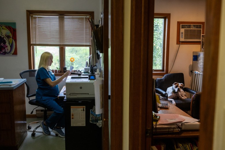 A person in blue scrubs looks at paper while seated at a desk in one room as a dog sits on a chair in an adjoining room.