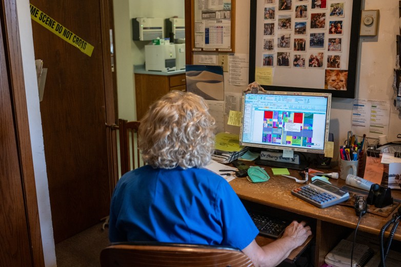 A person in blue scrubs sits at a desk facing a computer displaying a color-coded schedule, with papers, office supplies and photos nearby.