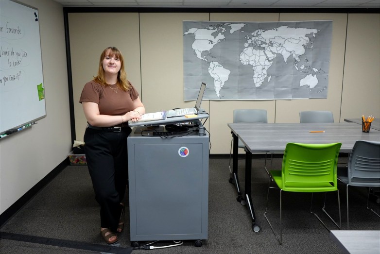 A person stands beside a laptop on a lectern in a room with a world map, a whiteboard, tables and green and gray chairs.