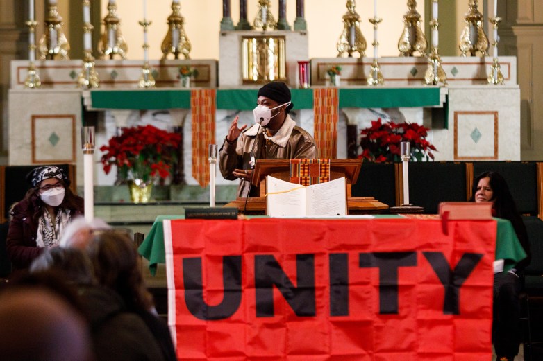 A person wearing a face mask stands and motions with hands at a lectern inside a church behind a large red banner reading