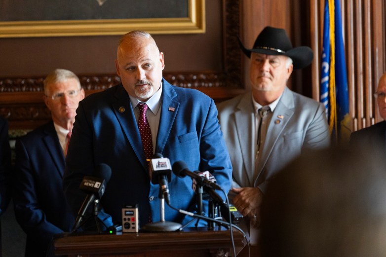 A person in a blue suit stands behind several microphones at a podium while people in suits stand behind, including one wearing a black cowboy hat.