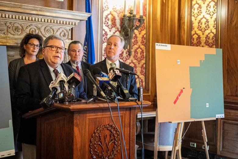 A person in a suit stands behind microphones at a wooden podium as three people stand behind and a color-coded map marked 