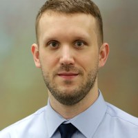 A person wearing a light blue collared shirt and dark tie looks toward the camera against a blurred background.