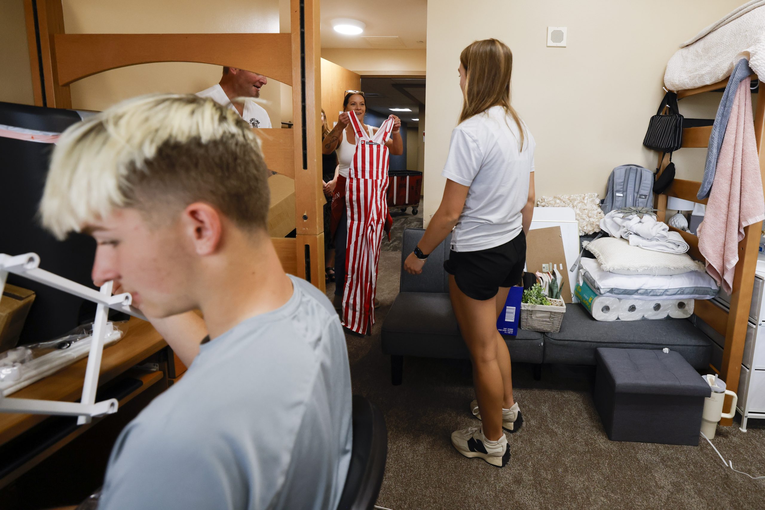 People stand in a room with lofted beds; one person holds red-and-white striped overalls.