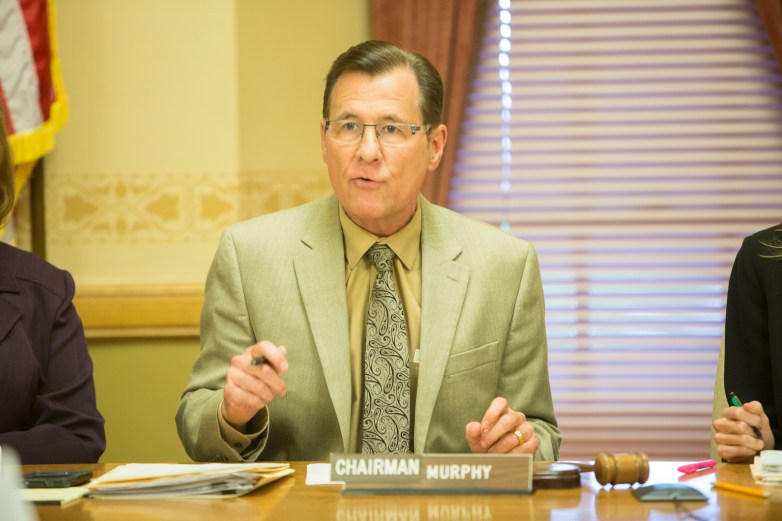 A person wearing glasses and a suit gestures at a table behind a nameplate reading 