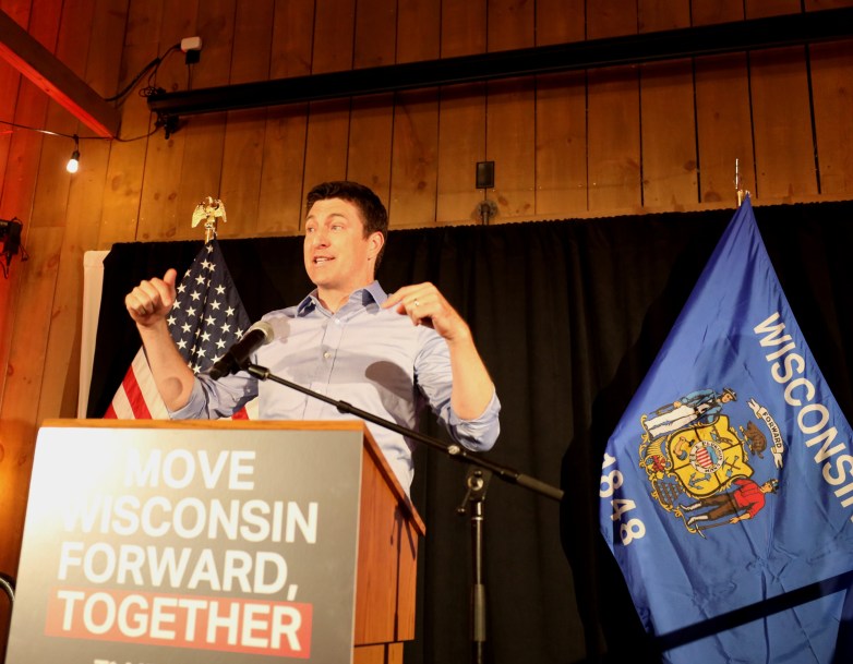 A person in a light blue shirt gestures with both hands while standing at a podium reading 
