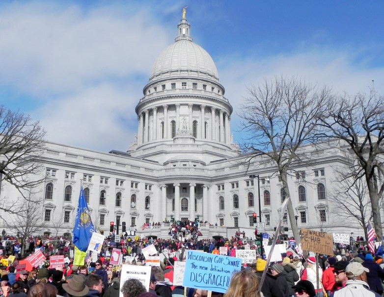 A large crowd holds signs and flags outside a domed government building beneath a blue sky.