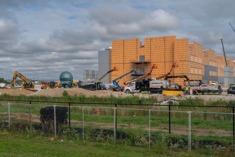 A large building with yellow exterior panels stands under construction, surrounded by excavators, boom lifts, trucks and people in safety vests behind a fence.
