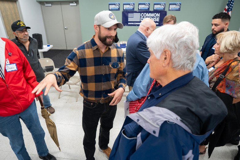 A person in a plaid shirt and gray cap gestures while standing with people in a room with 