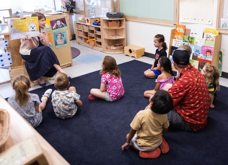 A person seated in a classroom holds up a picture book featuring a white dog while several people sit on a blue rug, surrounded by shelves of toys and books.
