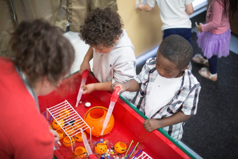 Two people use large plastic droppers at a red activity table filled with orange pumpkin containers, colorful beads and water as others stand nearby.