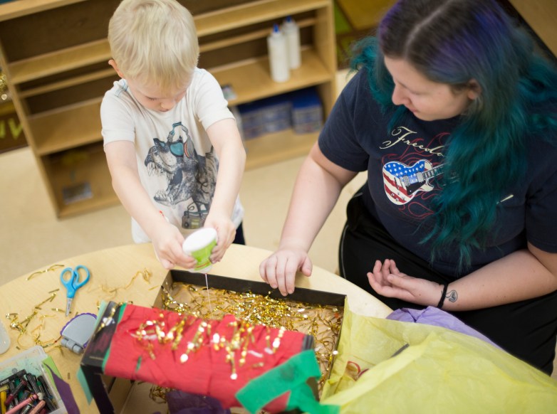 A person with blond hair squeezes glue onto gold tinsel inside a cardboard box while another person with blue and purple hair watches at a table covered with craft supplies.