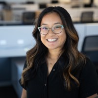 A person with long dark hair, glasses and a black buttoned shirt smiles at the camera in a room with desks and chairs in the background.